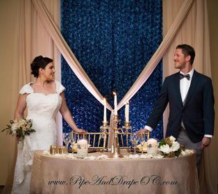 Bride and groom posing at sweetheart table in front of our gold and slate blue floral backdrop